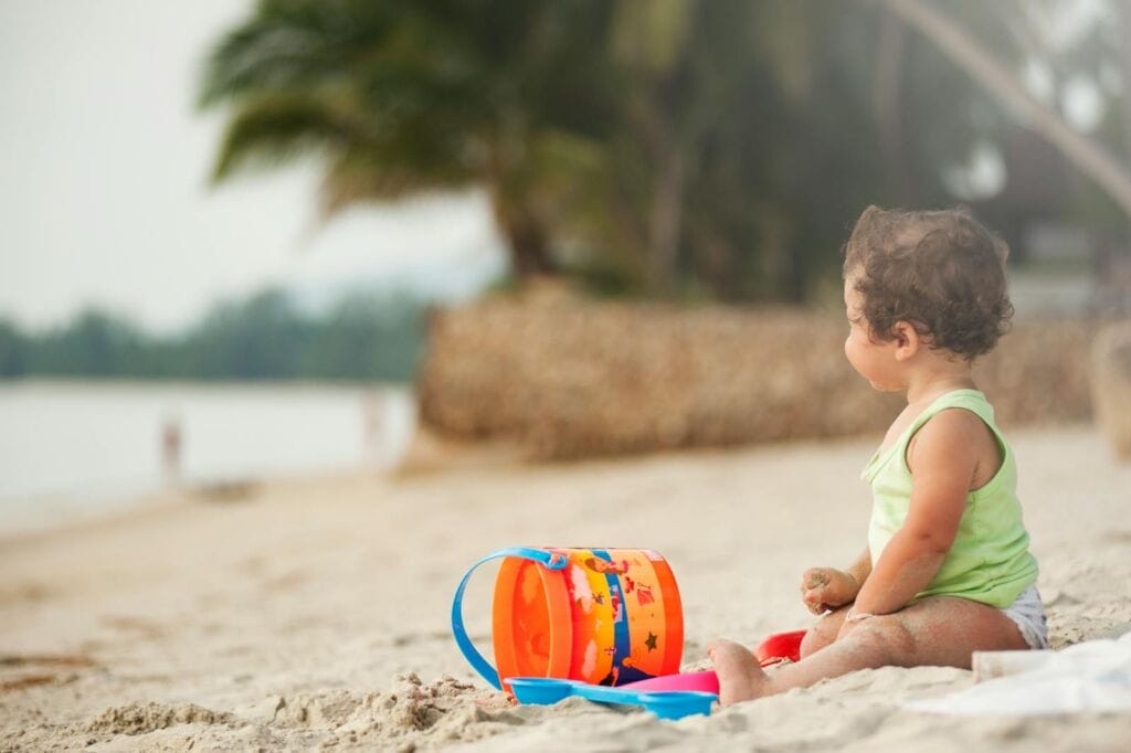 A young child plays happily with a bucket and sand on a tropical beach in Thailand.
